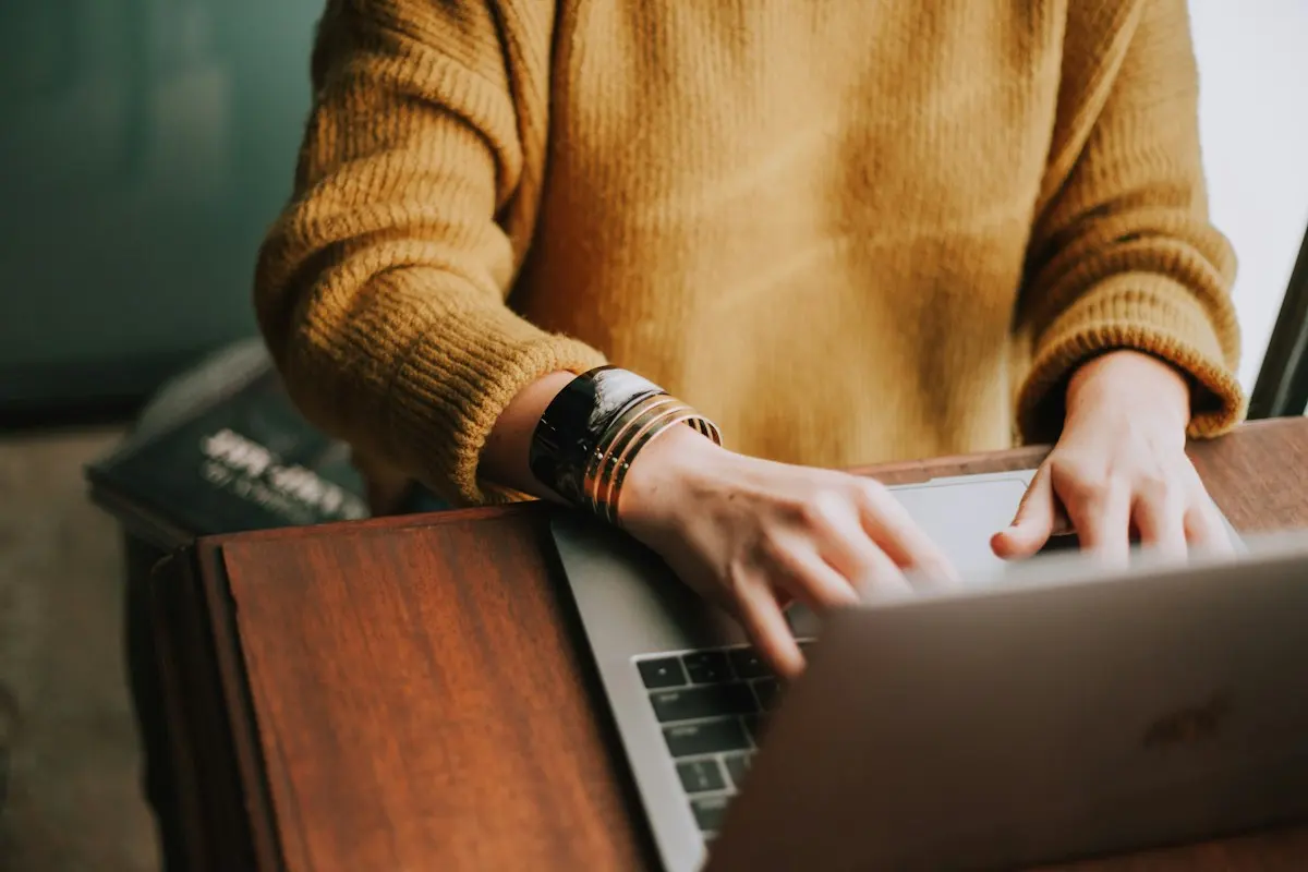 Someone in a mustard jumper typing on a laptop at a wooden table